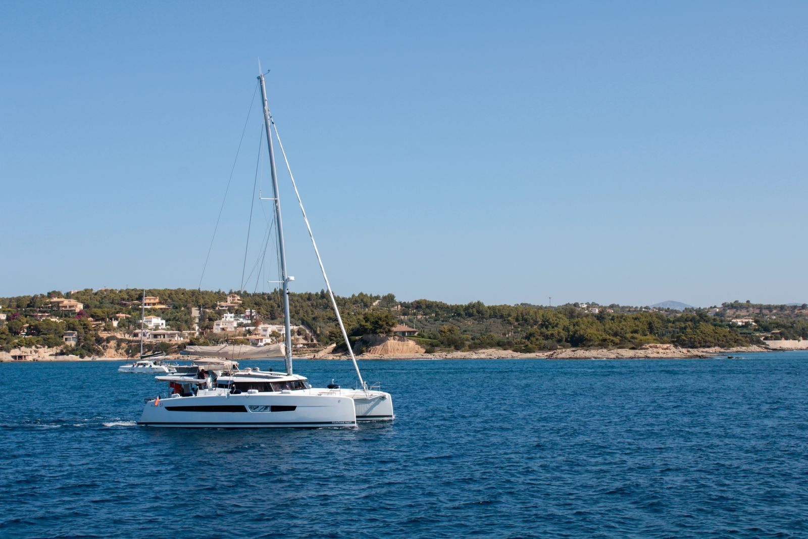 Sailing catamaran passing the green coastline of Hydra, a stop on a group sailing trip in Greece